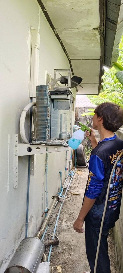 ช่างเตรียมล้างคอยล์ร้อนที่ติดผนังทางเดินข้างบ้าน โดยถือสายฉีดน้ำไว้