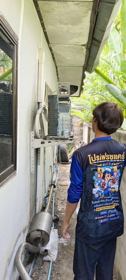 ช่างยืนถือสายฉีดน้ำเตรียมล้างคอยล์ร้อนบนผนังทางเดินแคบข้างบ้าน