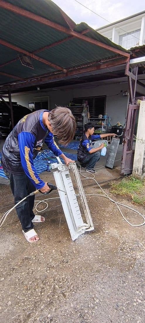 ช่างก้มฉีดน้ำล้างชิ้นส่วนแอร์บนลานปูนใต้ชายคาโรงจอดรถ