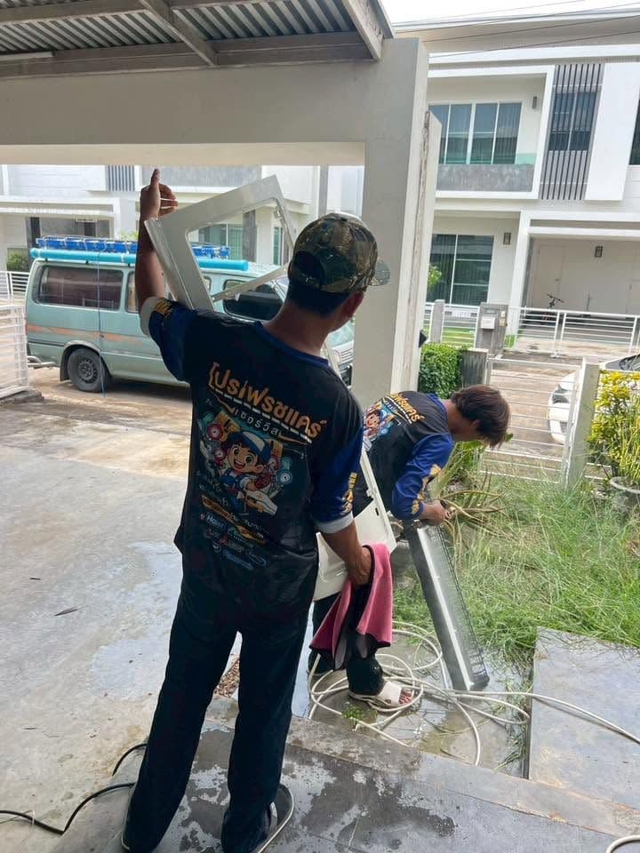 ช่างสองคนช่วยกันล้างชิ้นส่วนแอร์บนลานหน้าหมู่บ้านจัดสรร มีรถตู้จอดอยู่