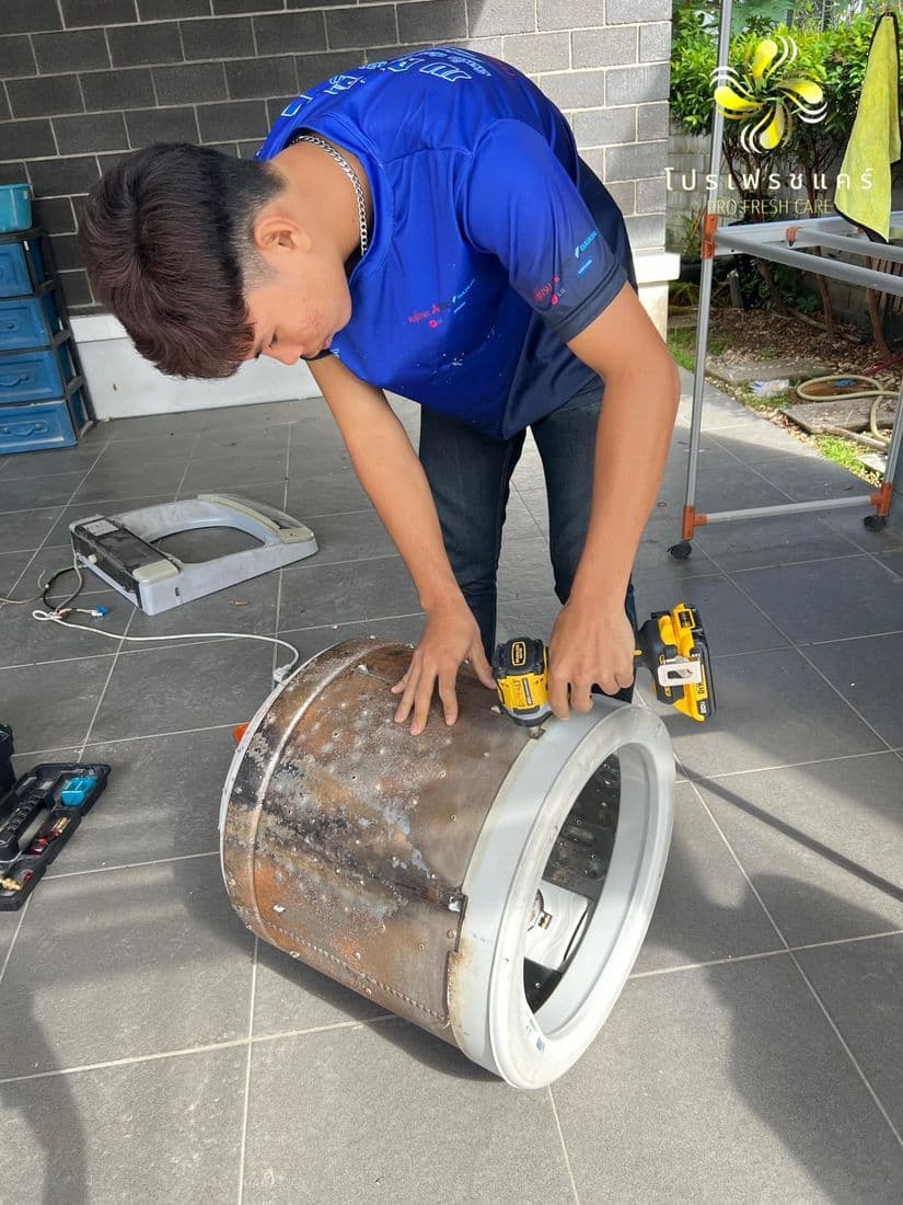 ช่างเสื้อฟ้าก้มขัดถังซักที่วางนอนกับพื้นกระเบื้อง มีคราบน้ำตาลติดรอบตัวถัง