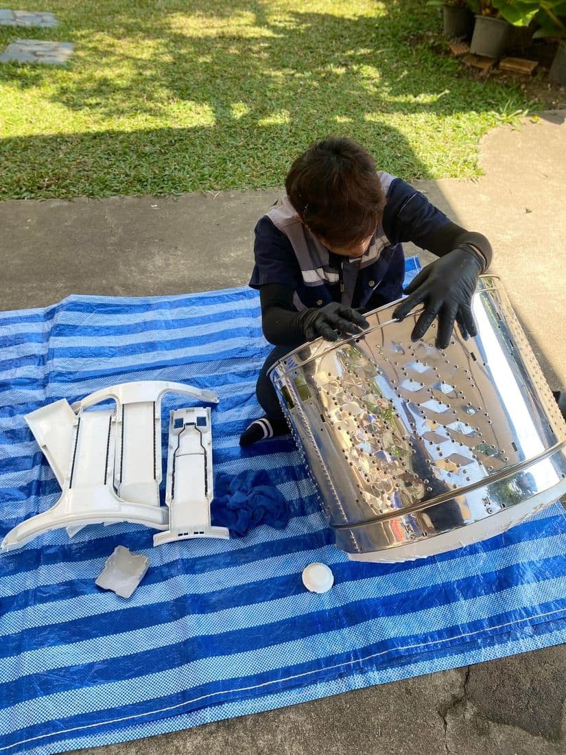 ช่างนั่งล้างถังบนผ้าใบที่ปูบนสนามหญ้า มีชิ้นส่วนพลาสติกวางเรียงรออยู่ข้าง