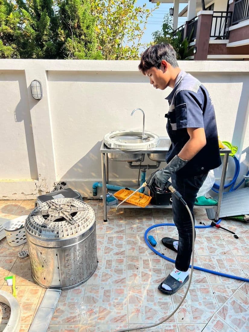 ช่างฉีดน้ำล้างชิ้นส่วนบนลานกระเบื้อง มีถังซักสองใบตั้งรออยู่คนละมุม