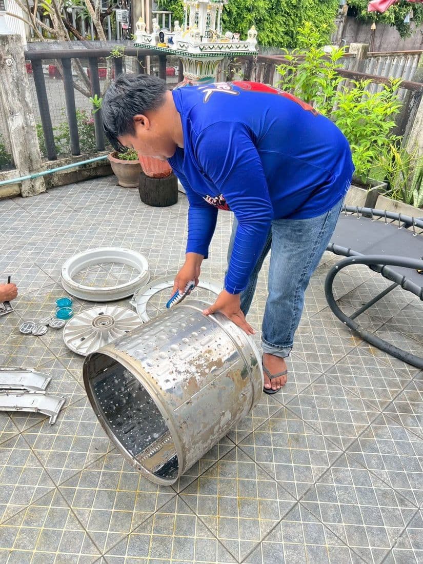 ช่างก้มยกถังซักขึ้นจากพื้น ข้างตัวมีสายฉีดน้ำและชิ้นส่วนวางเรียง