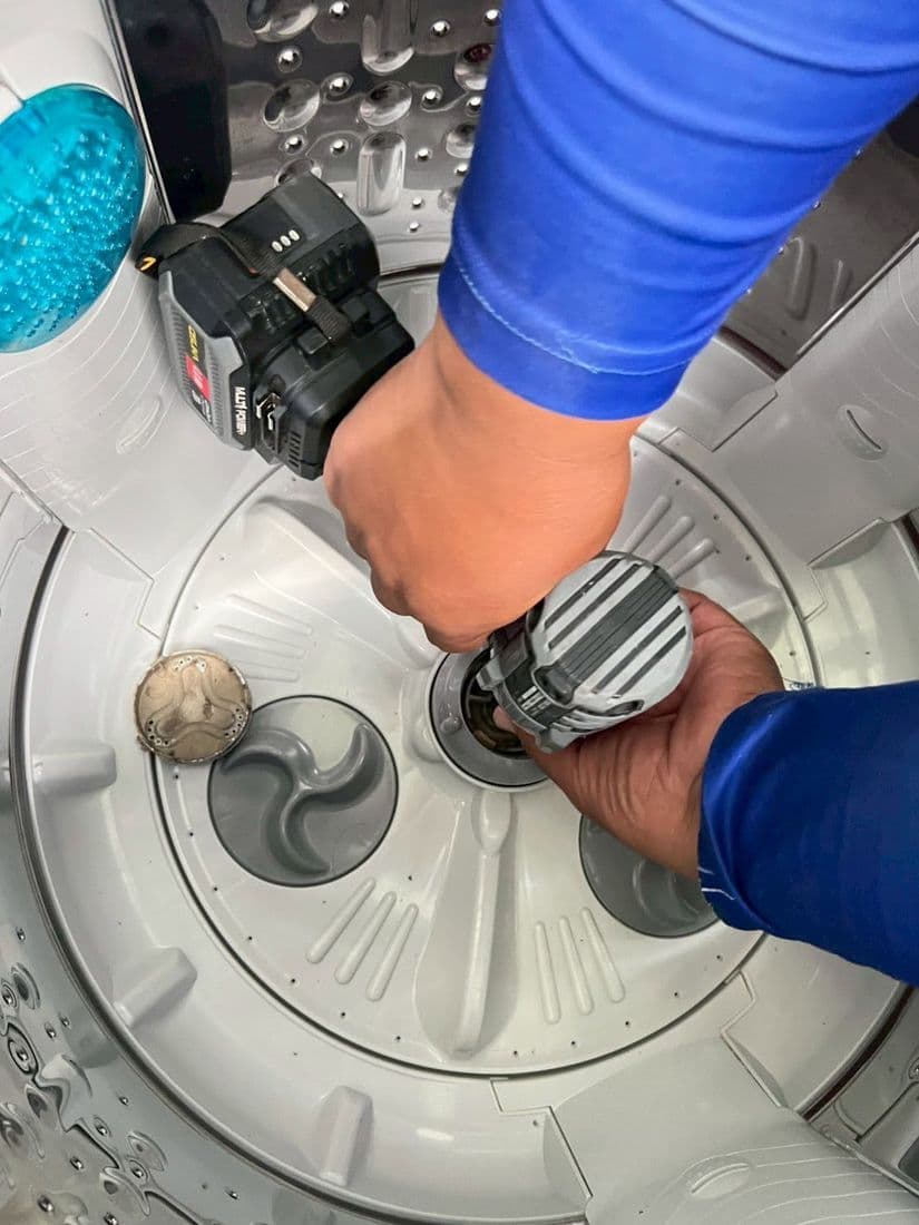 มือช่างใช้สว่านไฟฟ้าขันน็อตกลางถังเพื่อถอดจานซักออกจากแกน