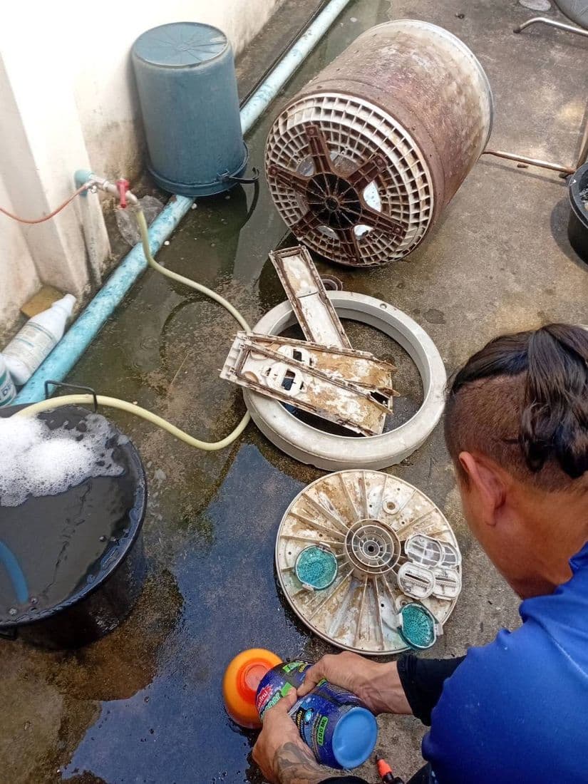 ช่างนั่งล้างชิ้นส่วนถังกับพื้นซีเมนต์เปียก ข้างเครื่องฉีดน้ำแรงดัน