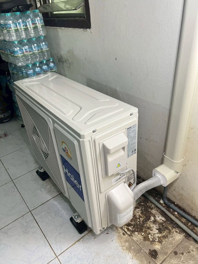 เครื่องแอร์ใหม่ยังห่อพลาสติกวางตั้งพิงมุมห้อง ข้างลังน้ำดื่มและแนวท่อ