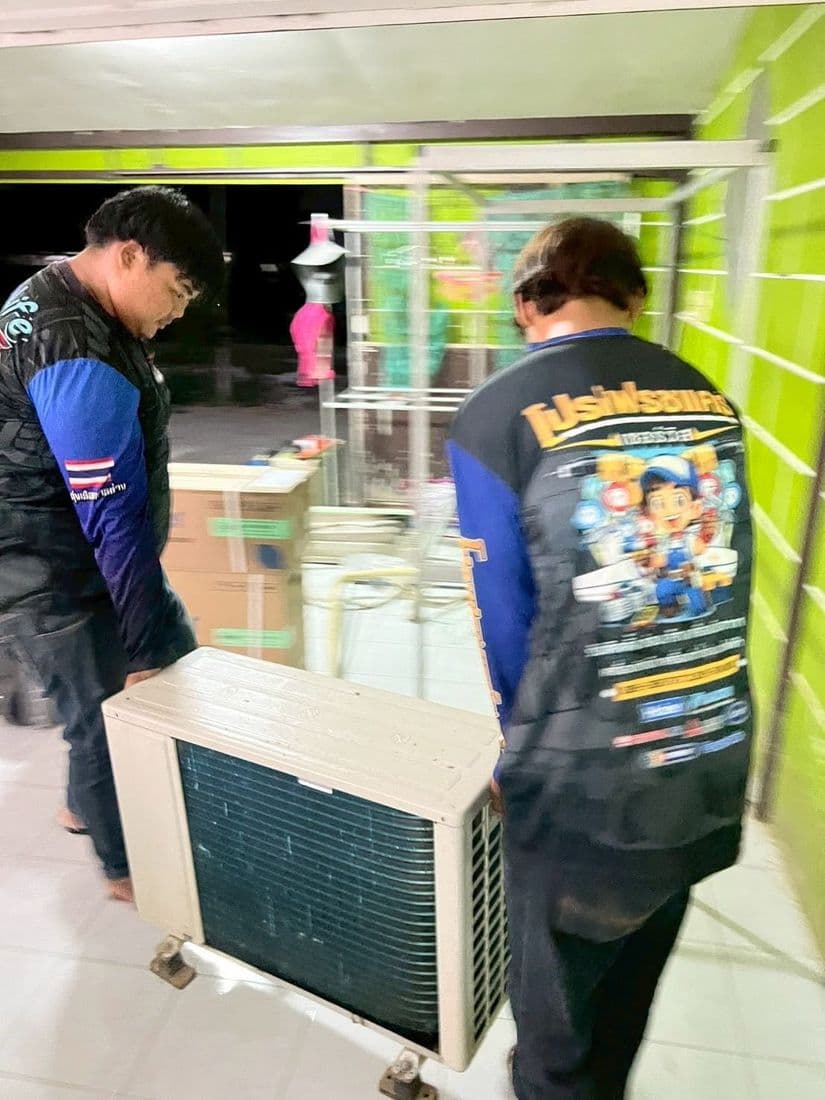 ช่างสองคนช่วยกันหามคอยล์ร้อนเครื่องเก่าผ่านหน้าร้านที่ผนังทาสีเขียว