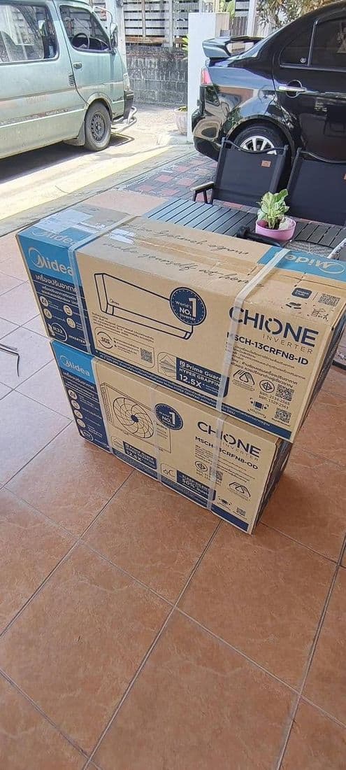 กล่องเครื่องปรับอากาศใหม่สองกล่องวางซ้อนบนพื้นกระเบื้องริมถนน ข้างรถที่จอดอยู่