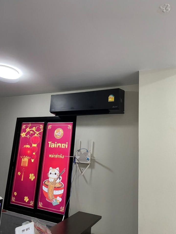 เครื่องปรับอากาศตัวสีดำติดผนังสูงในร้าน เหนือตู้แช่ที่มีป้ายสีชมพู