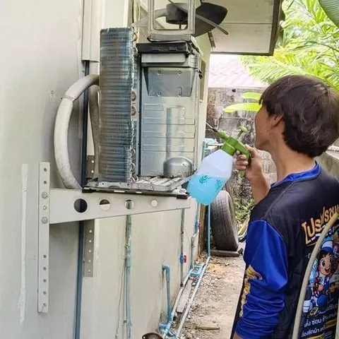 ช่างเตรียมล้างคอยล์ร้อนที่ติดผนังทางเดินข้างบ้าน โดยถือสายฉีดน้ำไว้