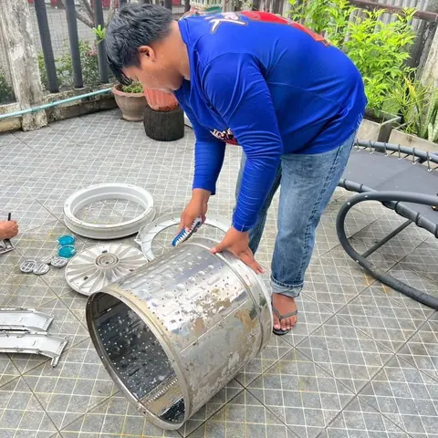 ช่างก้มยกถังซักขึ้นจากพื้น ข้างตัวมีสายฉีดน้ำและชิ้นส่วนวางเรียง
