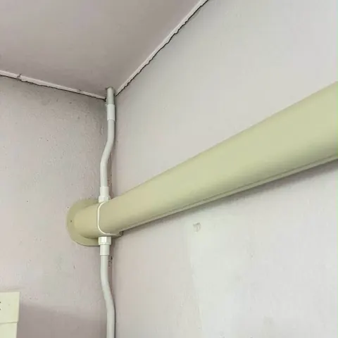 ท่อน้ำทิ้งสีขาวและรางครอบท่อสีครีมเดินคู่ขนานกันตามแนวมุมผนังห้อง
