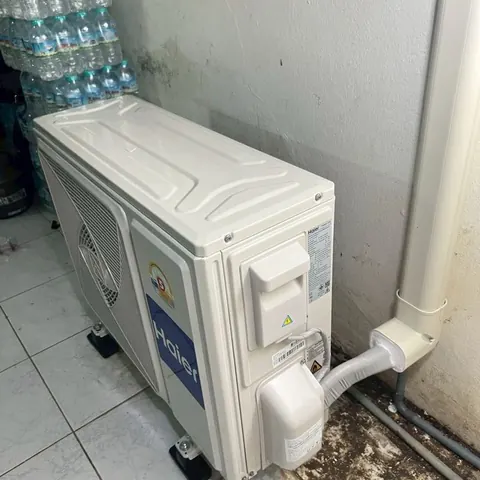 เครื่องแอร์ใหม่ยังห่อพลาสติกวางตั้งพิงมุมห้อง ข้างลังน้ำดื่มและแนวท่อ