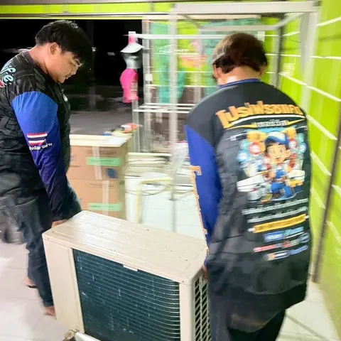 ช่างสองคนช่วยกันหามคอยล์ร้อนเครื่องเก่าผ่านหน้าร้านที่ผนังทาสีเขียว
