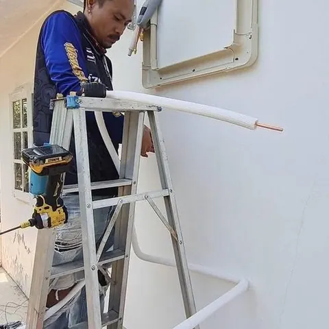 ช่างบนบันไดยึดขาเหล็กแขวนคอยล์ร้อน โดยมีท่อสีขาวพาดรออยู่